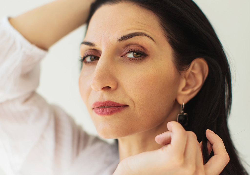 Confident woman with dark hair wearing earrings, looking directly at the camera while adjusting her hair.