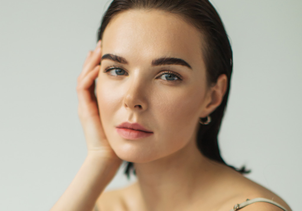 Close-up of a woman with clear skin and wet hair, softly resting her hand on her face while looking at the camera.