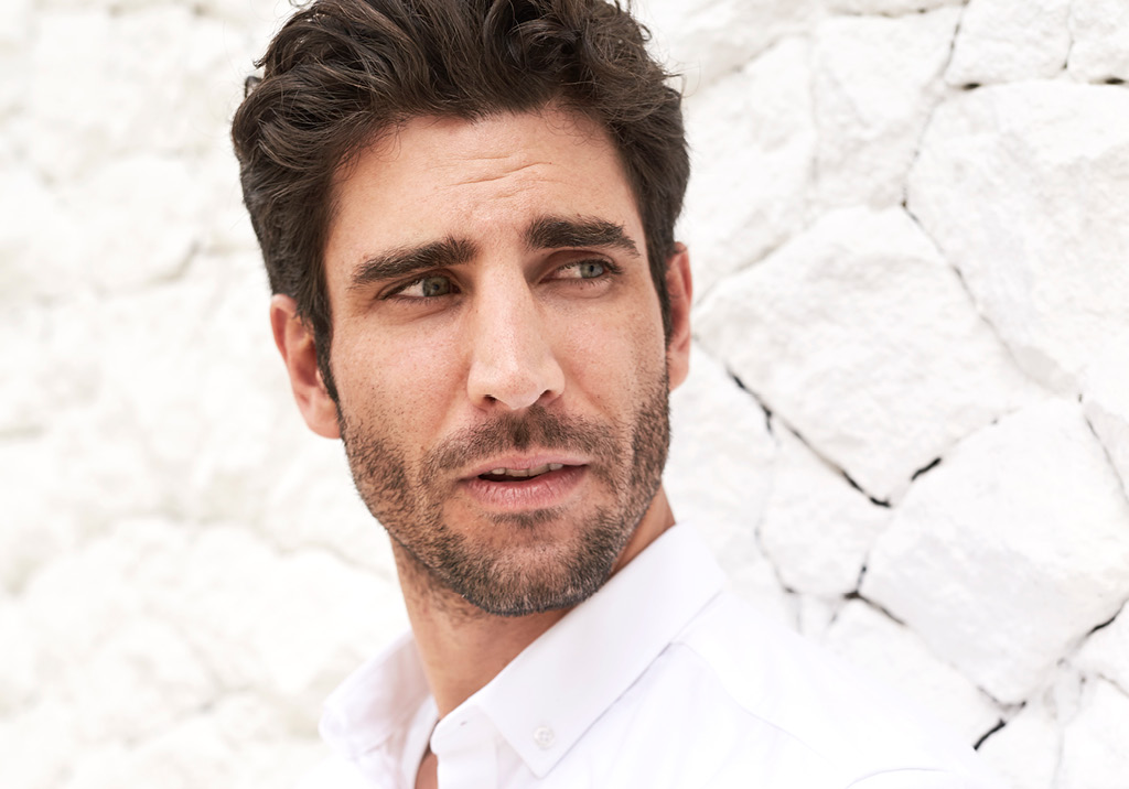 Man with dark hair and light stubble, wearing a white shirt, looking to the side against a white textured wall.