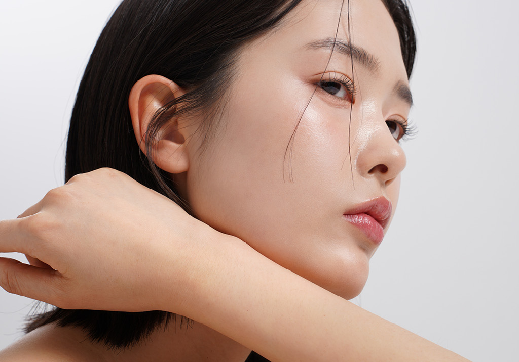 Side profile of a woman with smooth glowing skin, short dark hair, and a calm expression, hand near her neck.