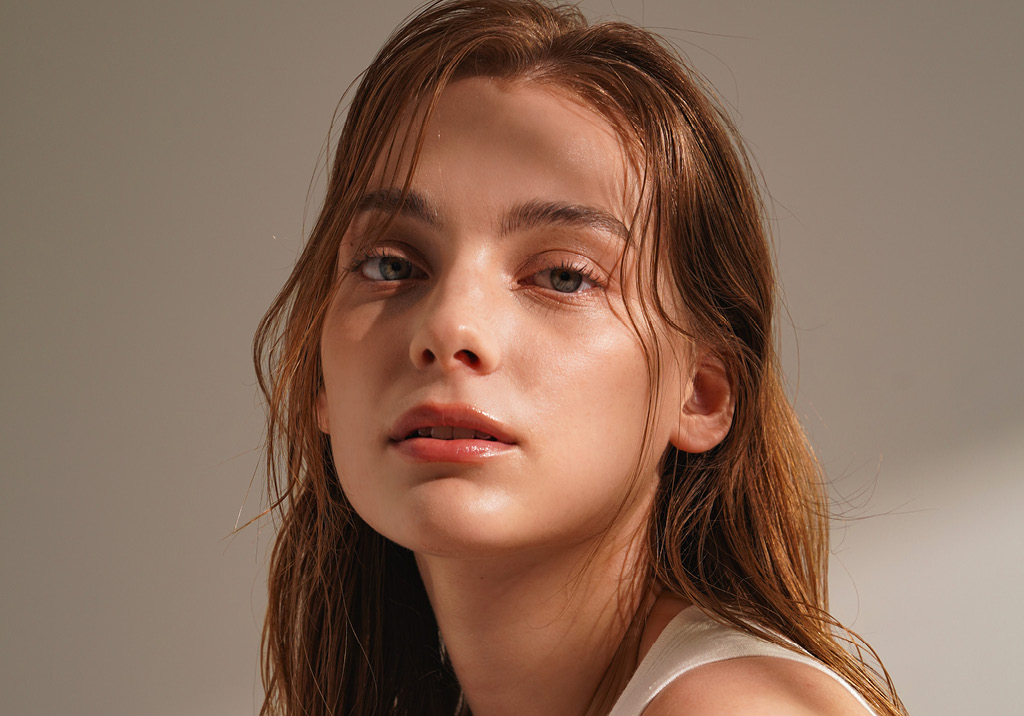 Close-up of a young woman with wet hair, natural makeup, and radiant skin, looking directly at the camera.