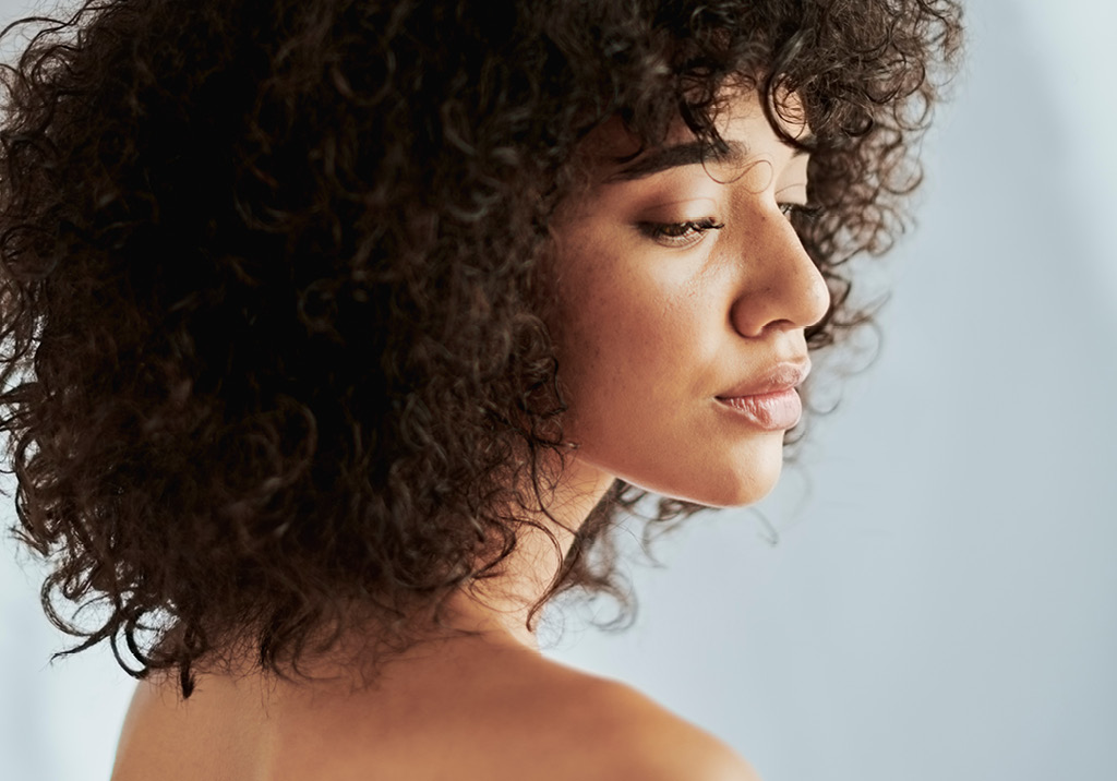 Side profile of a woman with voluminous curly hair, soft natural makeup, and serene expression against a light background.