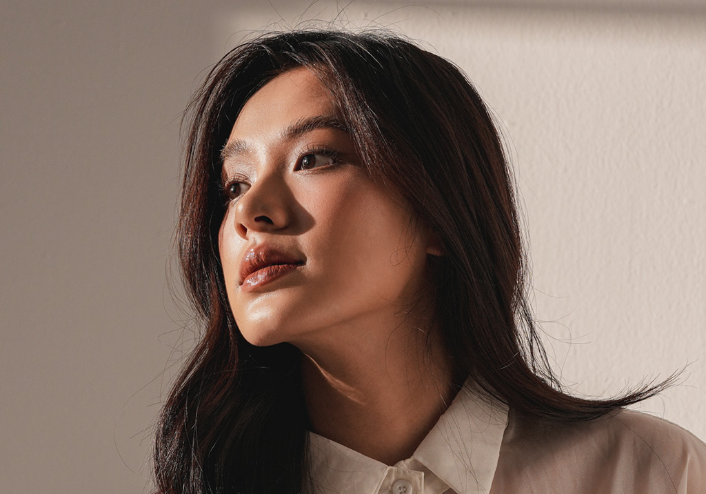 Woman with long dark hair wearing a white blouse, softly lit, gazing slightly upward with a calm expression.