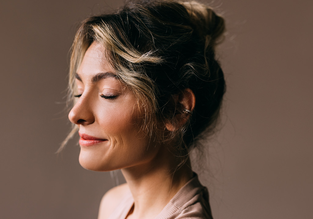 Smiling woman with light brown hair in a loose bun, eyes closed, and soft natural makeup against a warm background.