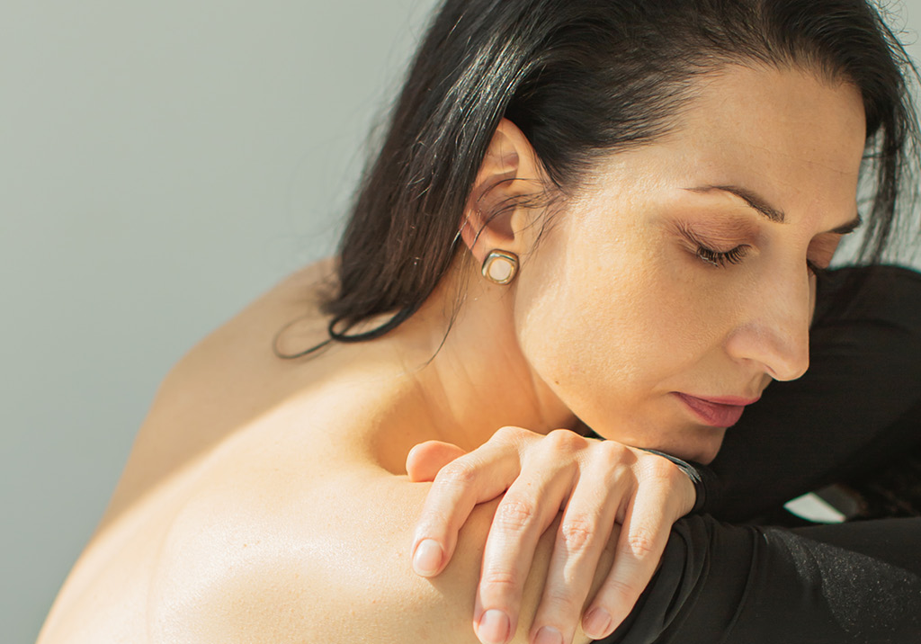 Woman with dark hair leaning forward, resting her arms on her bare shoulder, with eyes closed in soft light.