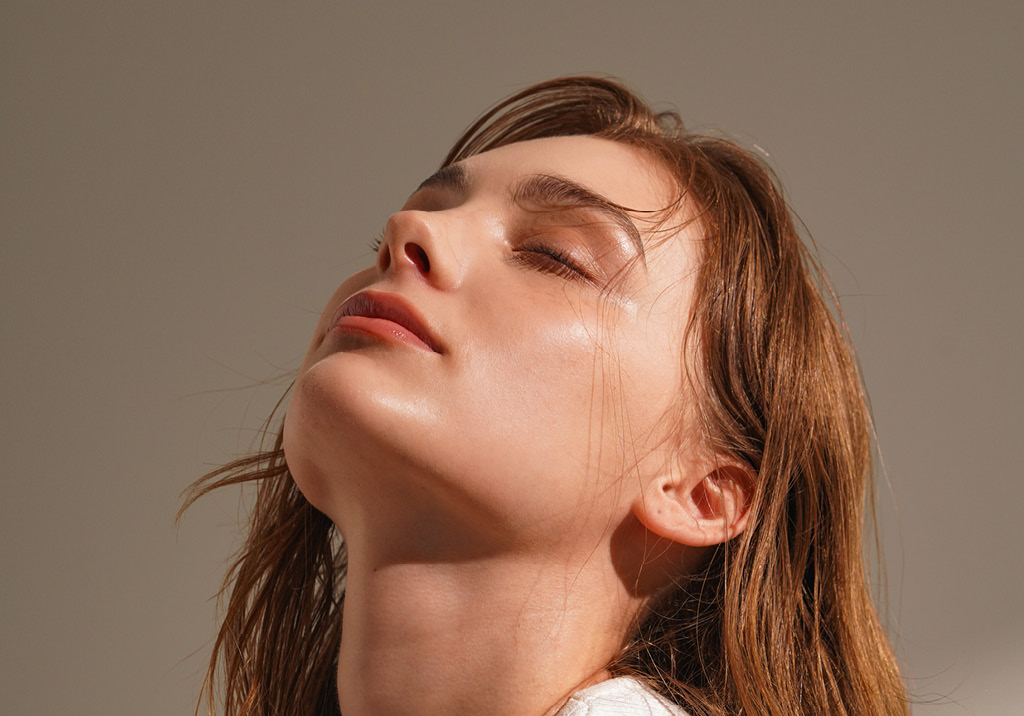 Young woman with wet hair tilting her head back slightly, eyes closed, glowing skin in warm sunlight.