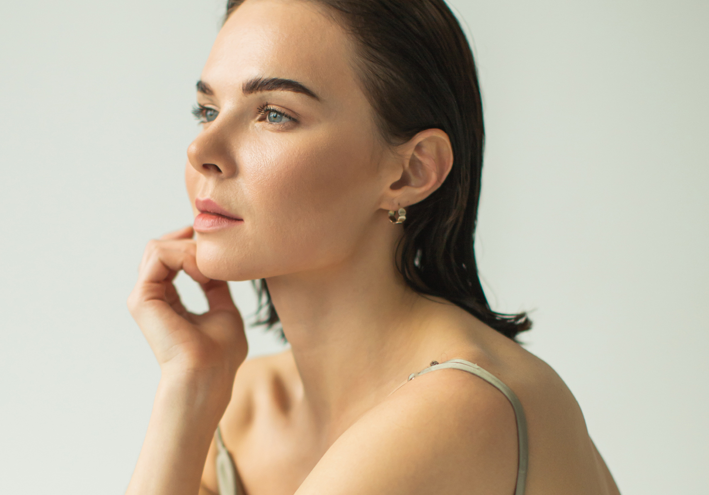 Woman with short dark hair looking thoughtfully to the side, resting her hand near her chin, wearing a sleeveless top and gold hoop earrings against a light background.